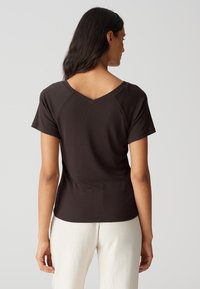 Dunkelbraunes Kurzarmhemd mit V-Ausschnitt und taillierter Passform. Glatte Textur mit Raglanärmeln und einem sauberen Abschluss am Saum.