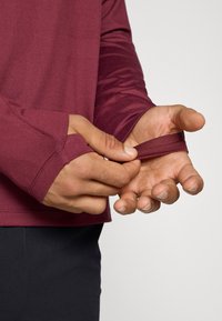 Maroon long-sleeve shirt with thumbholes, smooth texture, and fitted design, being adjusted at the cuffs. Black pants partially visible.