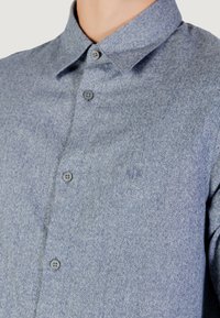 Camicia button-up in un tessuto blu con texture. Presenta un colletto classico, bottoni frontali e un piccolo logo vicino al petto.
