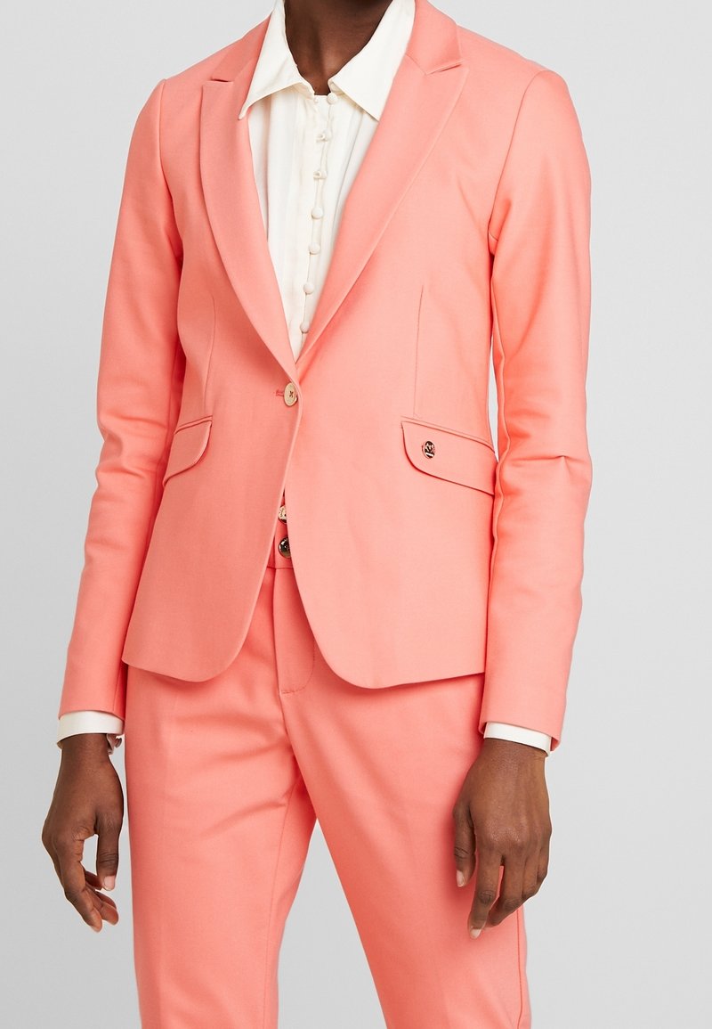 Femme portant un blazer rose corail ajusté et un pantalon assorti sur un chemisier blanc boutonné, debout devant un fond gris uni.
