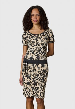 Kvinde med krøllet hår iført beige og sort kortærmet kjole med blomsterprint, sort talje og snøre, stående mod ensfarvet baggrund.