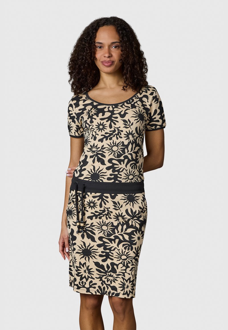 Frau mit lockigem Haar, die ein beige-schwarzes, kurzärmliges Kleid mit Blumenmuster, einem schwarzen Taillengürtel und Kordelzug trägt, steht vor einem schlichten Hintergrund.