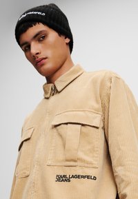 Beeži kootud jakk kahe eesmise taskuga, kaelusega ja musta "KARL LAGERFELD JEANS" logoga küljes. Must ribiga beanie.