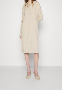 Beige stickad klänning med V-ringning, långa ärmar och vertikal ribbning, som sträcker sig till mitten av vaderna. Matchas med beige stilettklackar.