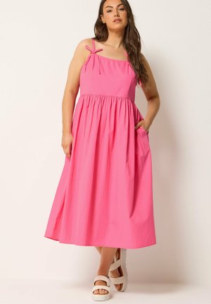 Kvinde iført en ærmeløs midi-kjole i pink med skulderbånd og hvide plateau-sandaler, stående mod en ensartet lys baggrund.