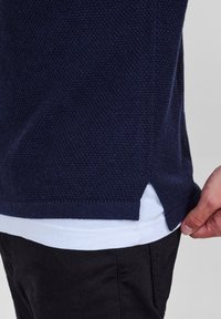 Marinblå stickad tröja i texturerat tyg, med sidöppningar, bärs över en vit T-shirt, med mörka byxor synliga längst ner.