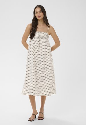 Žena v bielych midi šatách s čiernymi bodkami a tenkými ramienkami, skombinovaných s čiernymi sandálmi na podpätku s otvorenou špičkou, stojaca pred jednoduchým pozadím.