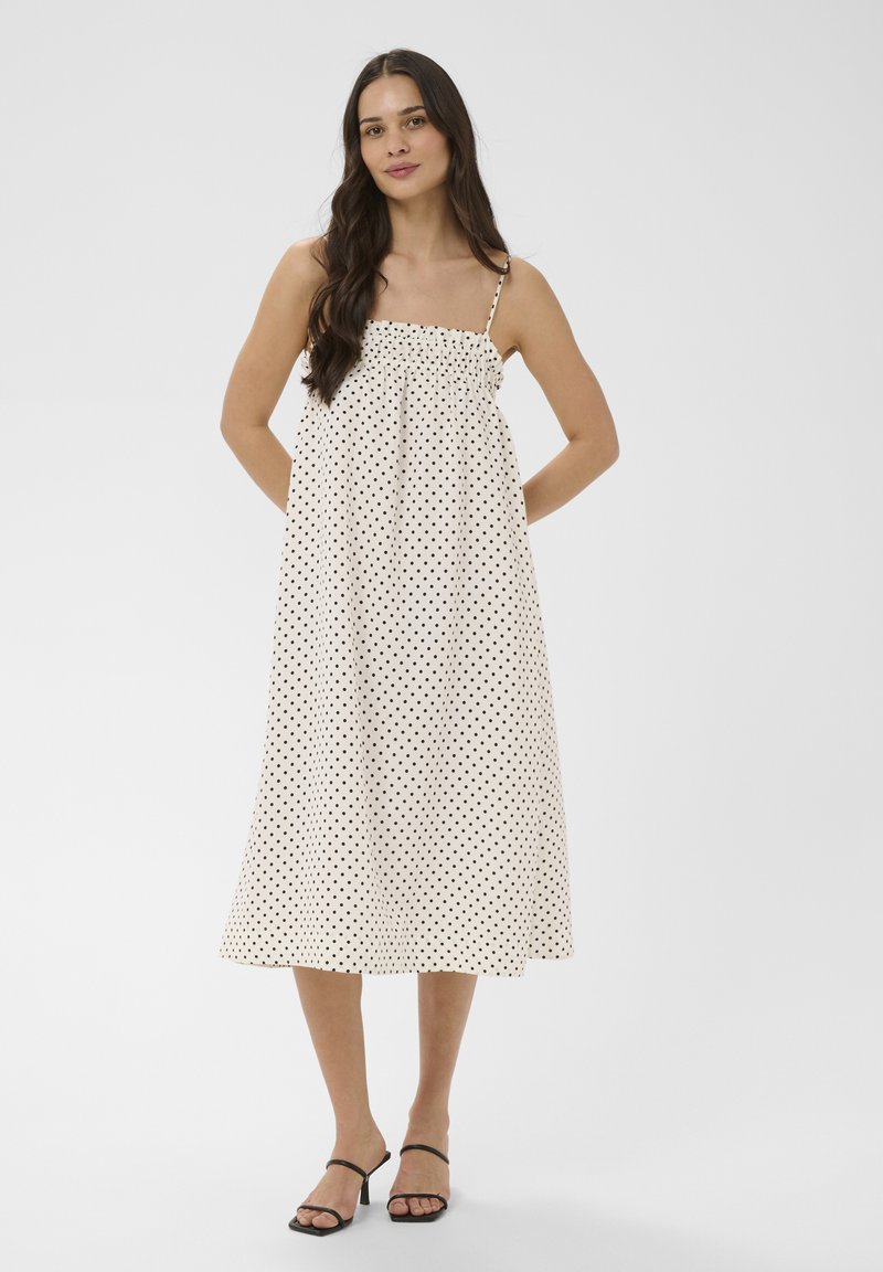 Femme portant une robe midi blanche à pois noirs avec fines bretelles, associée à des sandales noires à talons ouverts, debout devant un fond uni.