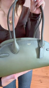 Zelena usnjena torbica z dvema ročajema, okrogle oblike, z zlatimi dodatki in pritrjeno oznako. Gladka tekstura z diskretnimi šivi.