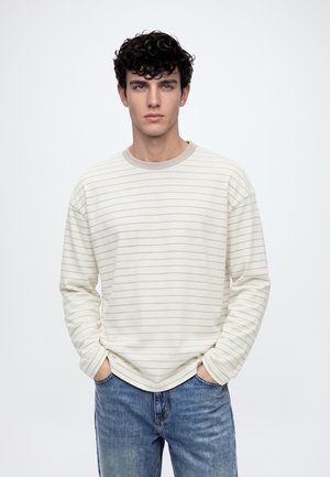 Jeune homme aux cheveux bouclés et foncés, portant une chemise crème à manches longues avec de fines rayures horizontales et un jean bleu, mains dans les poches.