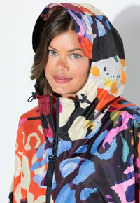Veste imperméable colorée avec un mélange de motifs abstraits en orange, noir et bleu. Comprend une capuche ajustable et une fermeture zippée.