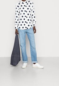 Osoba w białej koszuli z długim rękawem w czarne kropki, jasnoniebieskich, podwiniętych dżinsach, białych sneakersach, trzymająca dużą ciemnoniebieską torbę.