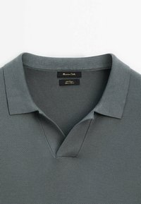 Polo en coton gris texturé avec col ouvert et étiquettes noires cousues à l'intérieur de l'encolure.