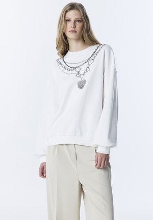 Frau mit langen blonden Haaren, die ein lockeres weißes Sweatshirt mit Ketten- und Herzdruck trägt, kombiniert mit beigefarbenen Hosen mit weitem Bein, stehend vor weißem Hintergrund.