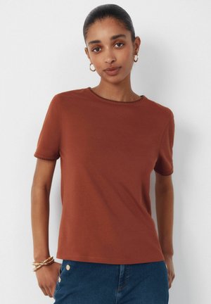 Junge Frau mit dunklen Haaren zu einem niedrigen Dutt gebunden, trägt goldene Creolen-Ohrringe, ein rostfarbenes kurzärmeliges Oberteil, blaue Jeans und goldene Armbänder.