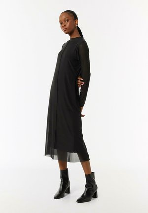 Zwarte jurk met lange doorschijnende mouwen en een rechte silhouet, voorzien van een split aan de voorkant. Gepaard met zwarte hoge enkellaarsjes.
