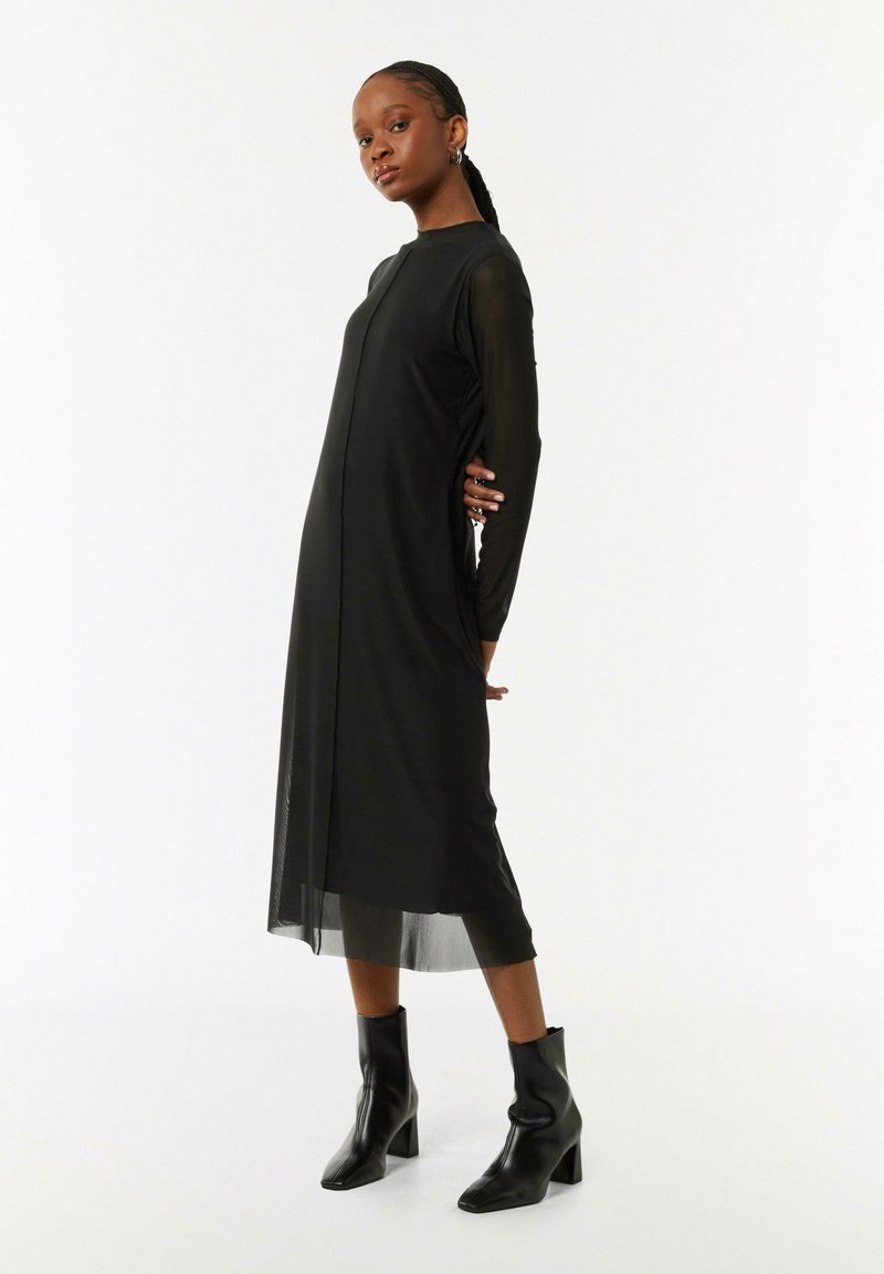 Schwarzes Kleid mit langen, durchsichtigen Ärmeln und geradem Schnitt, das einen vorderen Schlitz aufweist. Kombiniert mit schwarzen, hohen Stiefeletten.
