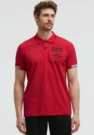 Mann trägt rotes kurzärmeliges Poloshirt mit schwarzem Text und Streifen an den Ärmeln, steht vor einem schlichten hellen Hintergrund.