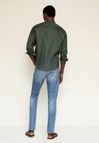 Camisa de manga larga de color verde oscuro con puños remangados, combinada con vaqueros azul claro. Modelo de espaldas, destacando un ajuste informal y un estilo relajado.