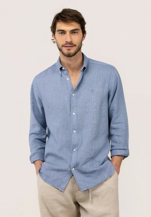 Hombre con cabello corto y castaño y barba, vestido con una camisa azul claro abotonada y pantalones beige con las manos en los bolsillos sobre un fondo liso.