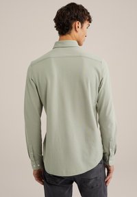 Cămiză lungă cu mânecă lungă de culoare verde deschis, cu guler, mansete cu nasturi și un tiv drept, fabricată dintr-un material neted cu o textură subtilă.