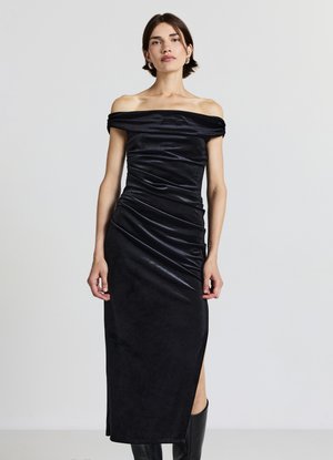 Abito nero scivolato sulle spalle con silhouette aderente, dettaglio arricciato, spacco laterale e tessuto liscio leggermente lucido. Lunghezza a metà polpaccio.