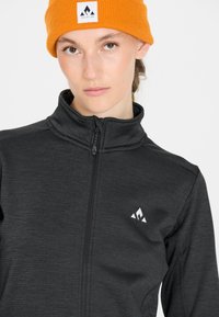 Svart zip-up jacka med hög krage, tillverkad av textil med struktur. Har en vit logotyp på vänster bröst och en stickad orange mössa.