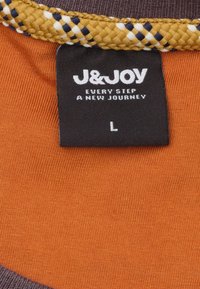 Bruin en oranje stof met een textuur geweven kraag. Het etiket toont "J&JOY," maat "L," en de zin "IEDERE STAP EEN NIEUWE REIS."