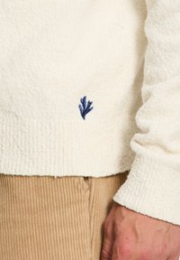 Biały sweter z miękkiego futerka o teksturowanym wykończeniu, z małym motywem niebieskiej koralowca na dole. Połączony z brązowymi spodniami z korduroyu.