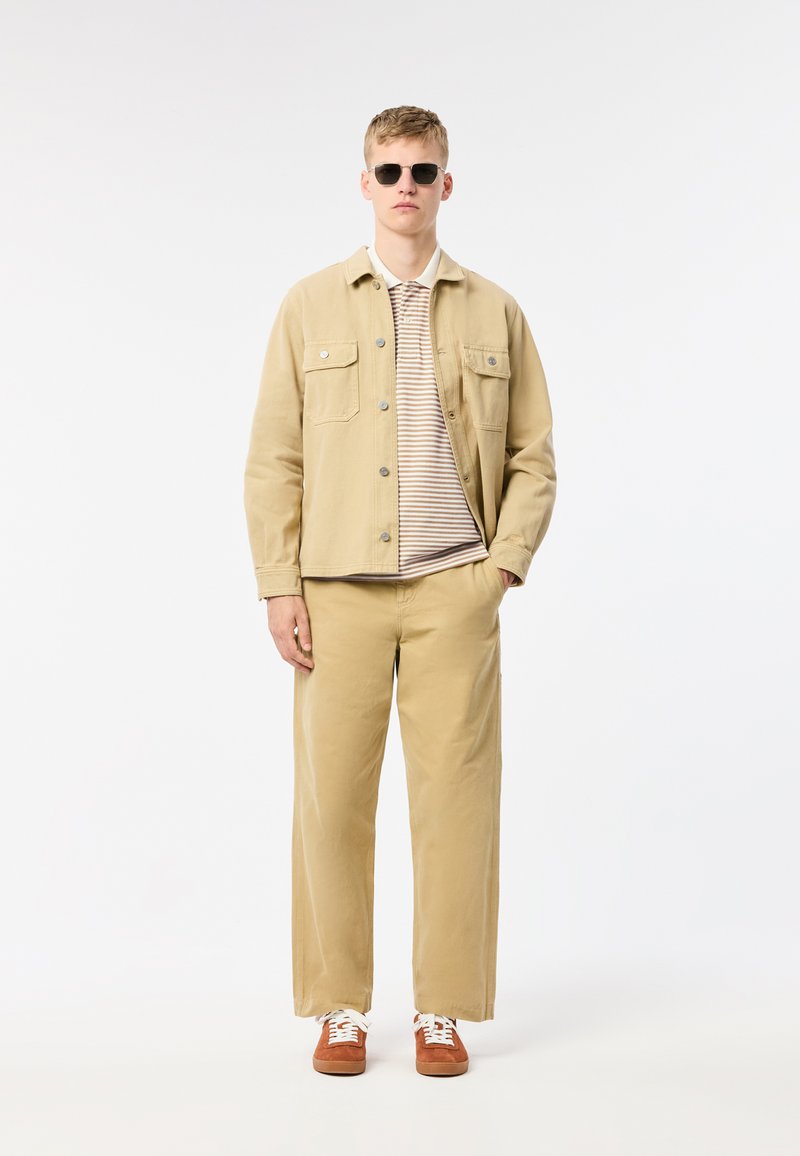 Veste en denim beige, polo rayé, pantalon large beige et baskets marron avec des lacets blancs. Mannequin portant des lunettes de soleil noires.