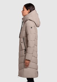 Vrouw in een lange beige gewatteerde jas met een hoge kraag en capuchon, staand in profiel tegen een effen achtergrond.