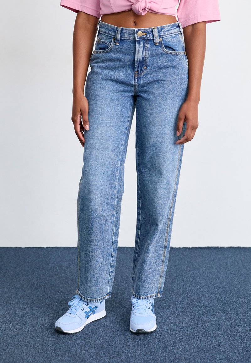 Woman wearing light blue wide-leg jeans, light blue sneakers, and a pink cropped t-shirt tied at the waist standing on dark carpet.