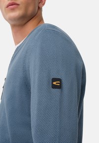 Blauer Strickpullover mit strukturiertem Stoff, gerippten Akzenten und einem Logo-Patch in Schwarz und Gold am linken Ärmel. Sichtbares Reißverschlussdetail.