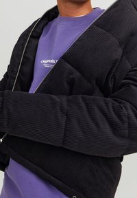 Czarna kurtka puchowa z sztruksu z zamkiem z przodu, charakteryzująca się prążkowaną fakturą i krótkim krojem, noszona na fioletowej bluzie.