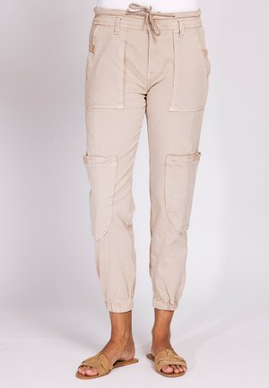 Beige Cargohose mit Kordelzugbund, großen Front- und Oberschenkeltaschen, elastischen Bündchen, getragen mit braunen Slides auf neutralem Hintergrund.