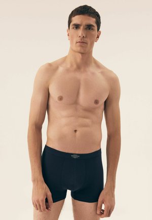 Black boxer shorts made of soft material, featuring a fitted design and a waistband with a brand label at the front, model standing in neutral light.