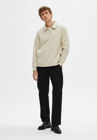 Pulover bej cu guler cu fermoar în treime, buzunar frontal și textură moale; asortat cu pantaloni negri cu picioare drepte și pantofi negri.