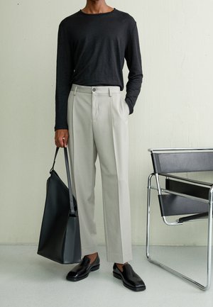 Homme portant une chemise noire à manches longues, pantalon gris clair ajusté, mocassins en cuir noirs, tenant un grand sac cabas noir, debout à côté d'une chaise noire.
