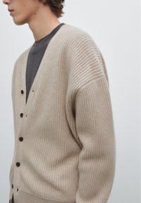Beige geribbelde cardigan met een V-hals, grote knopen en iets oversized mouwen, laag over een donkergrijze shirt.