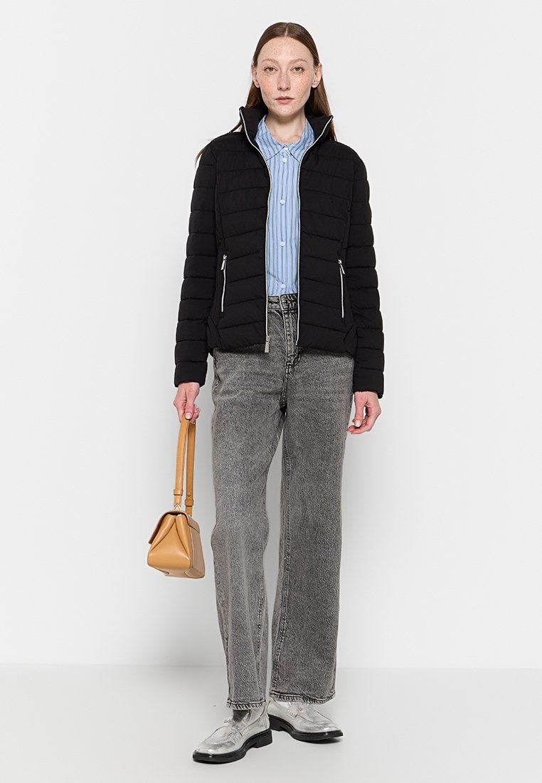 Giacca a vento nera con colletto alto, camicia a righe blu chiaro, jeans grigi, scarpe argentate e una piccola borsa beige.