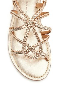 Sandalia de cuero dorado con tiras trenzadas formando un diseño circular en la parte superior. Textura suave con suela plana. Sin hardware visible.