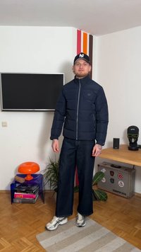Navy puffer jacket with a zippered front, paired with loose-fit dark denim pants and reflective sneakers. Room includes colorful decor.
