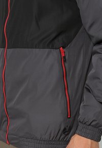 Giacca a vento grigia e nera con dettagli di cerniere rosse, polsini elastici e una tasca con cerniera sul lato sinistro.