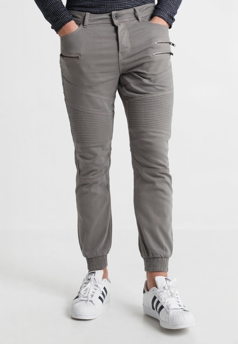 Graue elastische Hosen mit gerippten Nähten und Reißverschlusstaschen, kombiniert mit weißen Sneakers mit schwarzen Streifen.