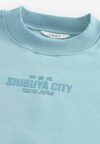 Svetlo modrý sweatshirt s rebrovaným okrúhlym výstrihom. Vyšívaný nápis znie "SHIBUYA CITY TOKYO JAPAN" v mierne vyvýšenom štýle.