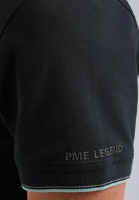Černé polo tričko s texturovanou látkou, s jemně vyšitým logem "PME LEGEND" na rukávu a kontrastním lemem na spodním okraji.