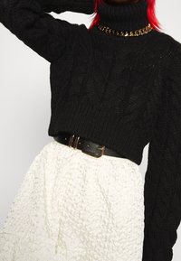 Czarny sweter z golfem w splot kablowy w połączeniu z białą spódnicą o strukturze, ściągnięty w talii czarnym paskiem z złotą klamrą.