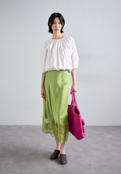 Hvid bluse med ballonærmer, grøn satin nederdel med blondekant, brune ribbede slip-on sko, pink vævet tote bag. Glatte teksturer og klare farver.