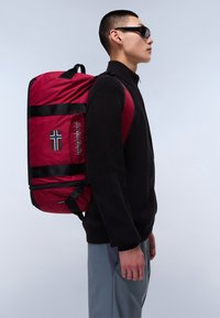 Punainen reppu mustilla hihnoilla, logolla ja Norjan lipun päällekkäin. Reppu on suorakulmainen, valmistettu kiiltävästä materiaalista ja sitä käytetään mustan suoran päällä.