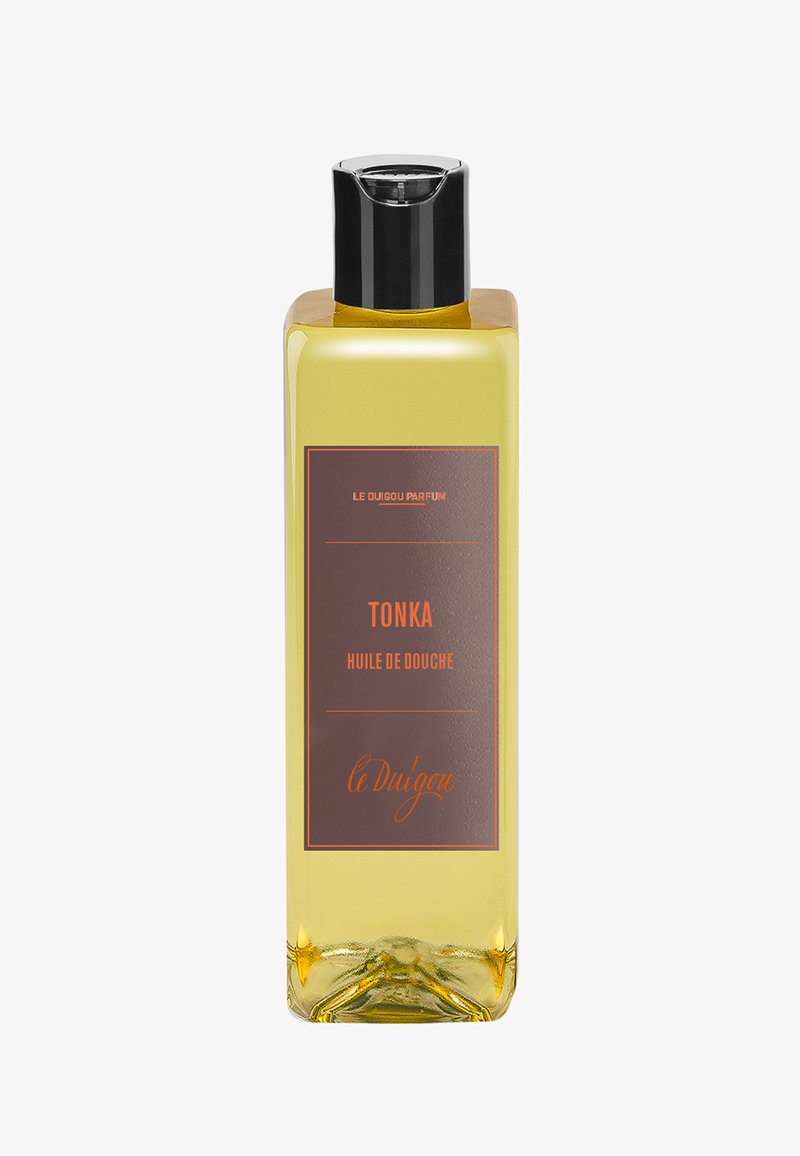 Klare rechteckige Flasche mit goldenem Duschöl und schwarzem Deckel. Das vordere Etikett zeigt einen braunen Hintergrund mit orangefarbener Schrift, die "Tonka Huile de Douche" besagt.